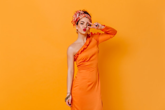 Coquettish woman in silk long dress and headband blows kiss and shows peace sign on orange space.