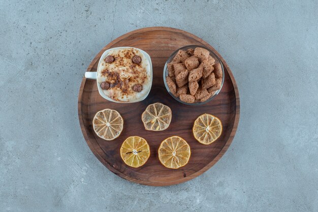Подушечки кукурузы на стакане рядом с ломтиками лимона и чашкой капучино на деревянной тарелке, на синем фоне. Фото высокого качества