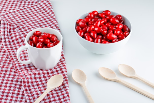 Cornel berries in bowl and cup with wooden spoons high angle view on picnic cloth and white