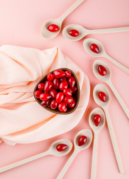 Cornel berries in wooden spoons and bowl on pink and textile, top view.