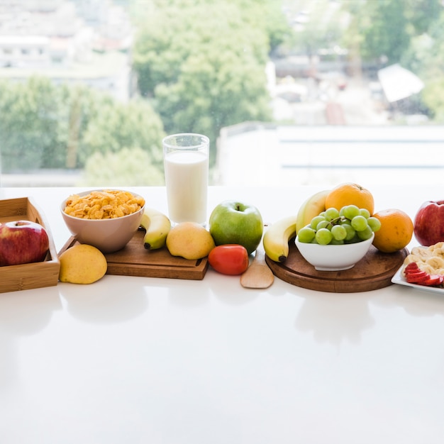 Cornflake bowl and colorful fruits with milk glass on white table near the window