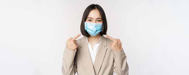 Coronavirus and business people concept Asian female entrepreneur pointing fingers at medical face mask at workplace standing over white background