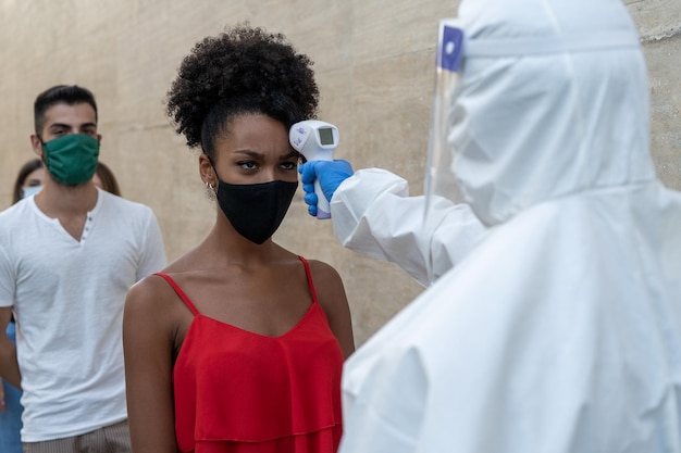 다른 젊은이들과 줄을 서 있는 아프리카계 여성의 적외선 디지털 온도계로 이마 온도를 측정하는 의사와 코로나바이러스 체크 포인트