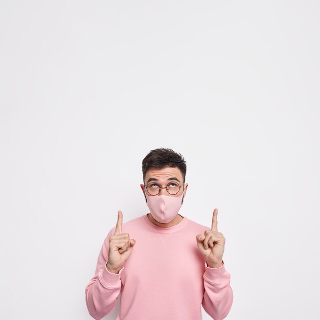 Coronavirus covid 19 concept. Young man wears hygienic mask to prevent contagious disease dessed in casual pink jumper has respiratory illness indicates upwards on blank space against white wall