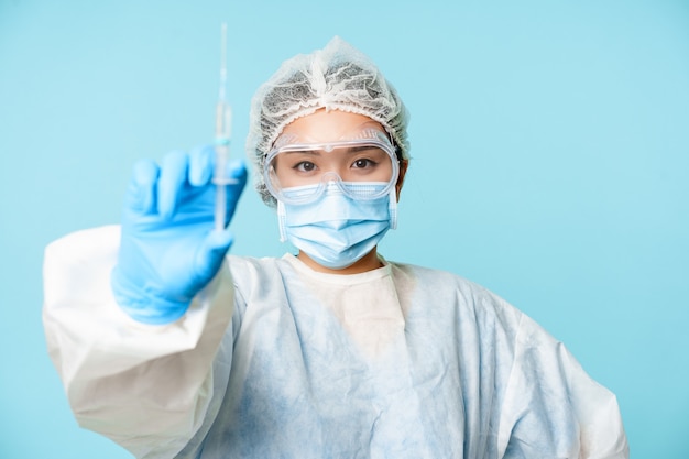 코로나바이러스와 의료 개념. 아시아 여성 의사 또는 간호사, covid-19, 독감 백신이 든 주사기를 보여주고 개인 보호 장비에 서서 파란색 배경