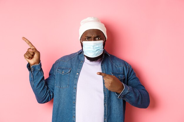 Коронавирус, образ жизни и концепция глобальной пандемии. Сердитый и разочарованный афро-американский мужчина в маске, указывающий влево, недовольно глядя в камеру, розовый фон.