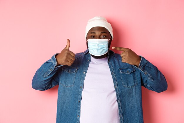 코로나바이러스, 라이프스타일 및 글로벌 전염병 개념. 젊은 아프리카계 미국인 남자는 얼굴 마스크를 가리키고 엄지손가락을 위로 올려 코비드, 분홍색 배경에서 자신을 보호합니다.