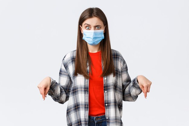 Coronavirus outbreak, leisure on quarantine, social distancing and emotions concept. Confused woman in medical mask cant understand what happening, pointing fingers down, look alarmed.