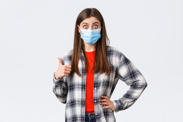 Coronavirus outbreak, leisure on quarantine, social distancing and emotions concept. Excited and surprised young woman in medical mask hear really good idea, show thumb-up in approval