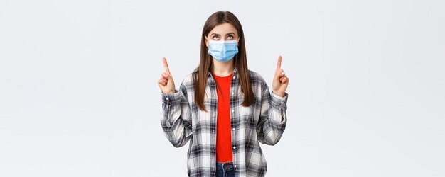 Досуг от вспышки коронавируса на карантине, концепция социального дистанцирования и эмоций Заинтересованная серьезная молодая женщина в медицинской маске и повседневной одежде смотрит и указывает на баннер