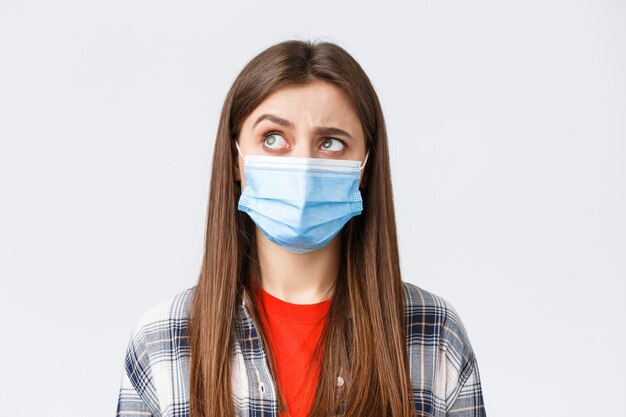 Coronavirus outbreak, leisure on quarantine, social distancing and emotions concept. Intrigued and hesitant young woman in medical mask, looking upper left corner reluctant, white background.