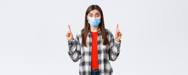 Coronavirus outbreak leisure on quarantine social distancing and emotions concept Surprised and astonished attractive woman in medical mask showing promo or link pointing fingers up