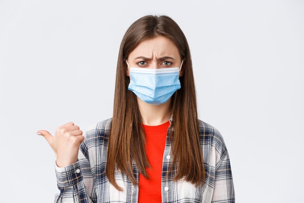 Coronavirus outbreak, leisure on quarantine, social distancing and emotions concept. Upset and disappointed young woman in medical mask frowning, condemn smth, pointing finger left displeased