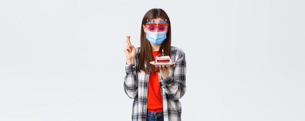 Coronavirus outbreak lifestyle social distancing and celebration concept Enthusiastic happy girl in glasses and medical mask celebrate birthday on quarantine cross fingers make wish at cake