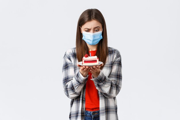 Coronavirus outbreak, lifestyle during social distancing and holidays celebration concept. Confused woman in medical mask staring puzzled at birthday cake lit candle, thinking, white background.