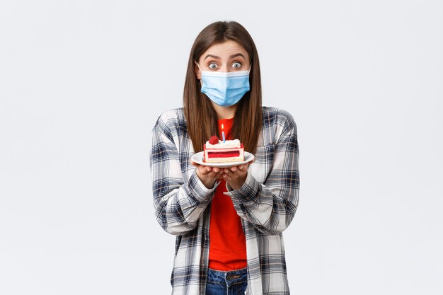 Coronavirus outbreak, lifestyle during social distancing and holidays celebration concept. Cute happy birthday girl making wish, wear medical mask, holding b-day cake, celebrating inside home.