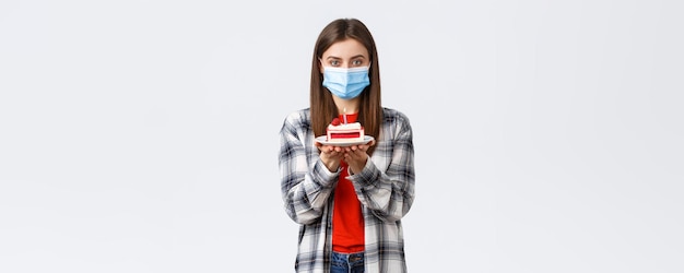 Coronavirus outbreak lifestyle during social distancing and holidays celebration concept Cute young woman in medical mask celebrating birthday alone during quarantine hold bday cake