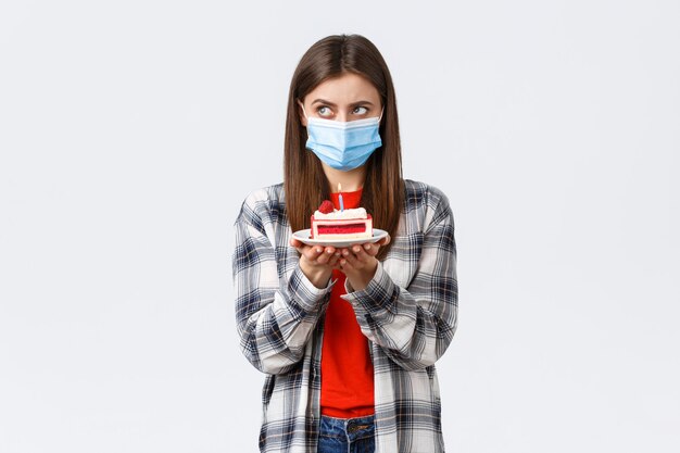 Coronavirus outbreak, lifestyle during social distancing and holidays celebration concept. Serious birthday girl in medical mask, focus on making wish, thinking as hold b-day cake with lit candle.