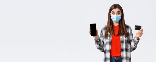 Coronavirus outbreak working from home online shopping and contactless payment concept Excited woman in medical mask showing credit card and mobile phone look surprised