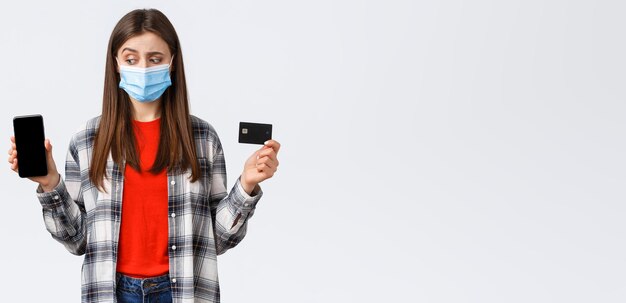 Coronavirus outbreak working from home online shopping and contactless payment concept Unsure girl in medical mask look as making decision at mobile phone show credit card and smartphone