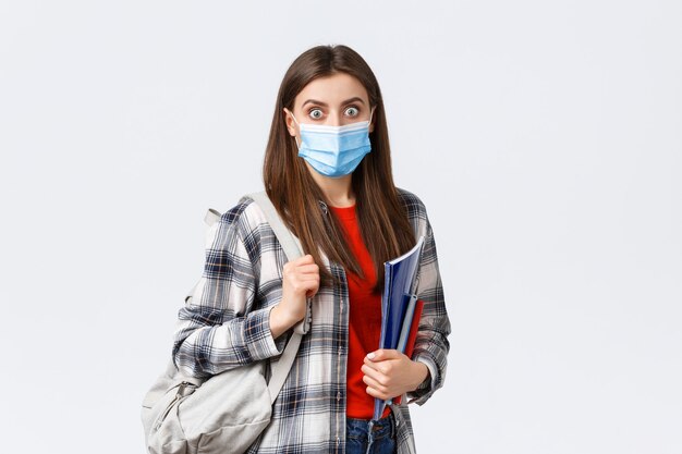 コロナウイルスのパンデミック、covid-19教育、そして学校に戻るという概念。医療マスクでショックを受けて驚いた女の子、学生はキャンパスで大きなニュースをあえぎ、ノートとバックパックを持っています