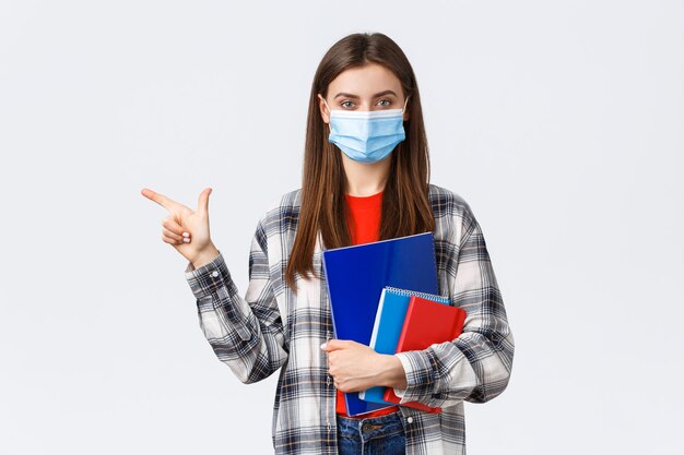 Coronavirus pandemic, covid-19 education, and back to school concept. Young pretty female student in medical mask with notebooks, pointing finger left, showing university info.