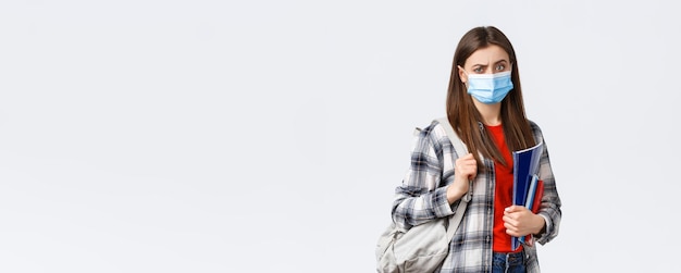 Coronavirus pandemic covid19 education and back to school concept Doubtful and skeptical female student in medical mask frowning disappointed hold notebooks and backpack