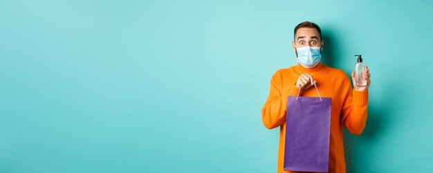 Coronavirus pandemic and lifestyle concept man in face mask showing shopping bag and hand sanitizer