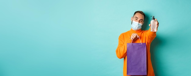 Free photo coronavirus pandemic and lifestyle concept man in face mask showing shopping bag and hand sanitizer