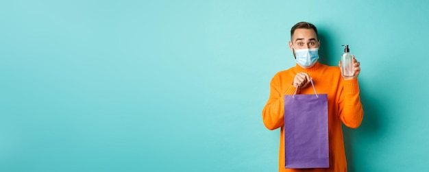 Free photo coronavirus pandemic and lifestyle concept man in face mask showing shopping bag and hand sanitizer