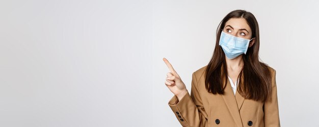 Free photo coronavirus and people concept portrait of business woman at workplace wearing face mask pointing fi