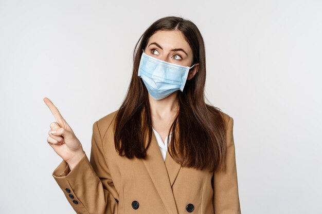 Coronavirus and people concept portrait of business woman at workplace wearing face mask pointing fi...