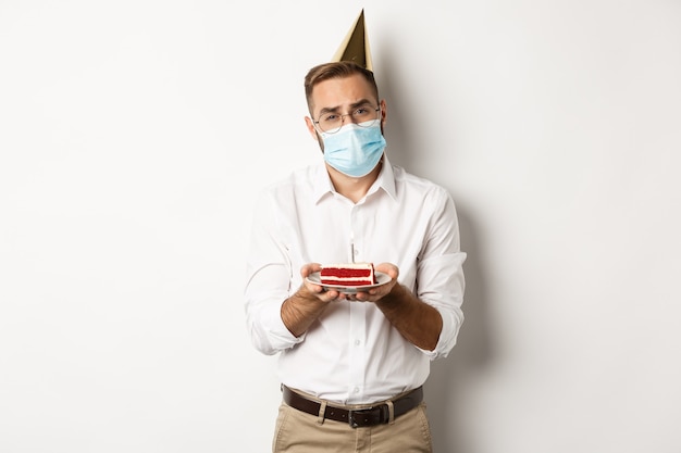 Coronavirus, quarantine and holidays. Sad man cant blow candle off birthday cake, wearing face mask and complaining, standing  