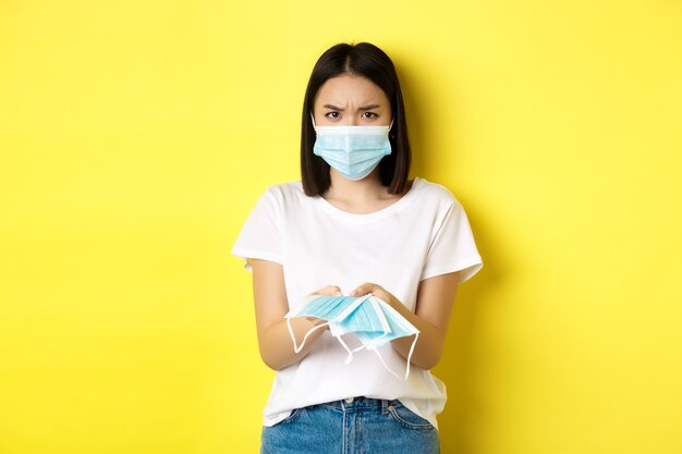 コロナウイルス、検疫および医学の概念。あなたに屋内で医療マスクを与える怒っているアジアの女の子、眉をひそめている、黄色の背景の上に立っている