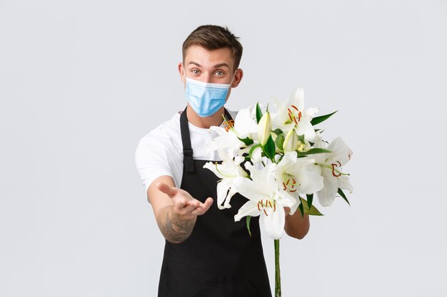 Coronavirus, social distancing business during covid-19 pandemic concept. Smiling handsome florist, salesman in flower shop wearing medical mask, reaching hand for payment and holding bouquet