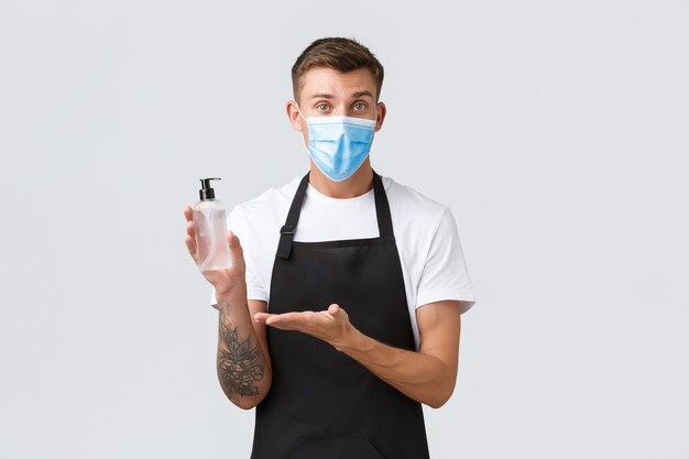 コロナウイルス、カフェやレストランでの社会的距離、パンデミックの概念の間のビジネス。陽気なセールスマンの医療用マスク、バリスタ、またはウェイターが、消毒のための手指消毒剤の重要性を説明します