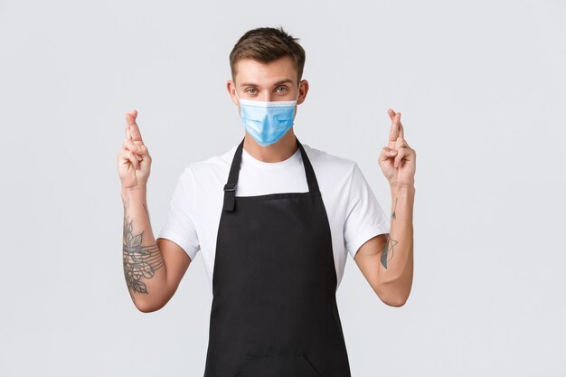 Coronavirus, social distancing in cafes and restaurants, business during pandemic concept. Confident hopeful barista, salesman in medical mask cross fingers good luck, making wish or pleading