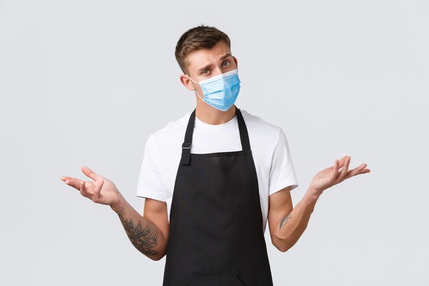 Coronavirus, social distancing in cafes and restaurants, business during pandemic concept. Confused handsome barista, salesman in medical mask shrugging, looking indecisive white background