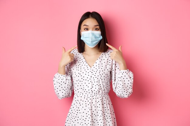 Coronavirus social distancing and lifestyle concept cute asian woman pointing at face mask asking to...