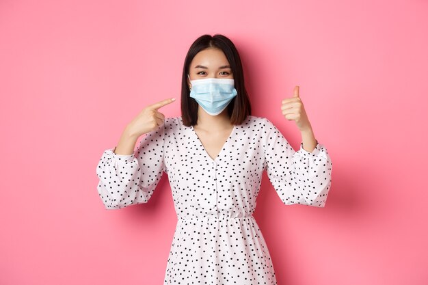 Coronavirus social distancing and lifestyle concept cute asian woman pointing at face mask showing t...