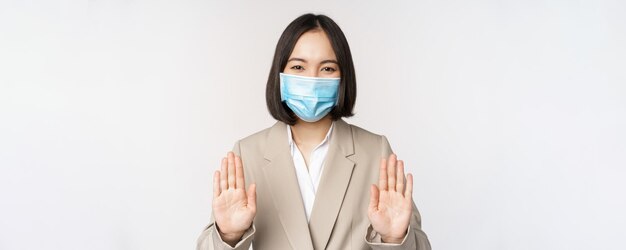 Coronavirus and work concept Portrait of asian female office lady woman at workplace wearing medical face mask and showing stop prohibition gesture white background