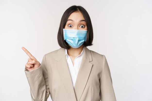 Coronavirus and work concept Portrait of woman in medical face mask pointing finger left showing logo or banner advertisement white background