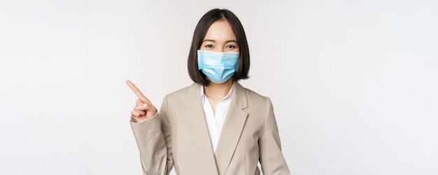Coronavirus e concetto di lavoro ritratto di donna con maschera medica che punta il dito a sinistra mostrando logo o banner pubblicitario sfondo bianco