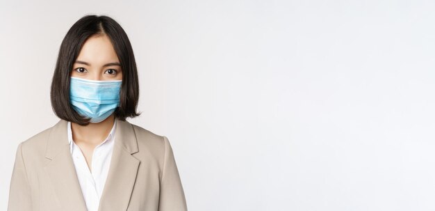 Coronavirus and workplace concept Portrait of asian office worker businesswoman in medical face mask looking at camera white background