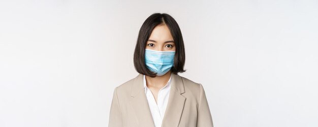 Coronavirus and workplace concept Portrait of asian office worker businesswoman in medical face mask looking at camera white background