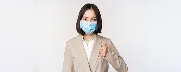 Coronavirus and workplace concept portrait of businesswoman in medical face mask showing thumbs up w