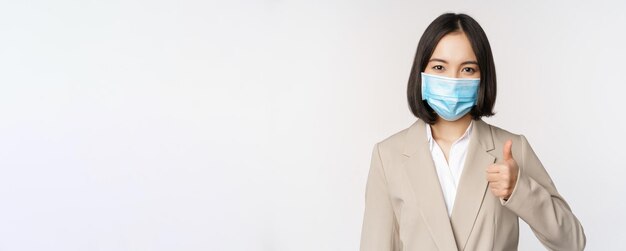 Coronavirus and workplace concept portrait of businesswoman in medical face mask showing thumbs up w