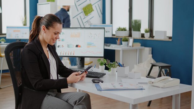 Corporate employee looking at smartphone screen while working on business planning with computer and charts at company office. Businesswoman using mobile phone for brainstorming ideas