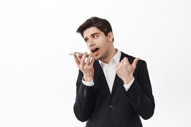 Corporate man talking speakerphone and counting on fingers give instructions during phone call standing with smartphone against white background
