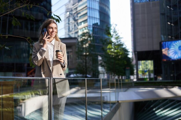 ベージュのスーツを着た企業の女性は、朝のコーヒーを飲み、スマートフォンの画面を見て、オフィスの外の通りに電話スタンドでメッセージをチェックするアプリケーションを使用しています